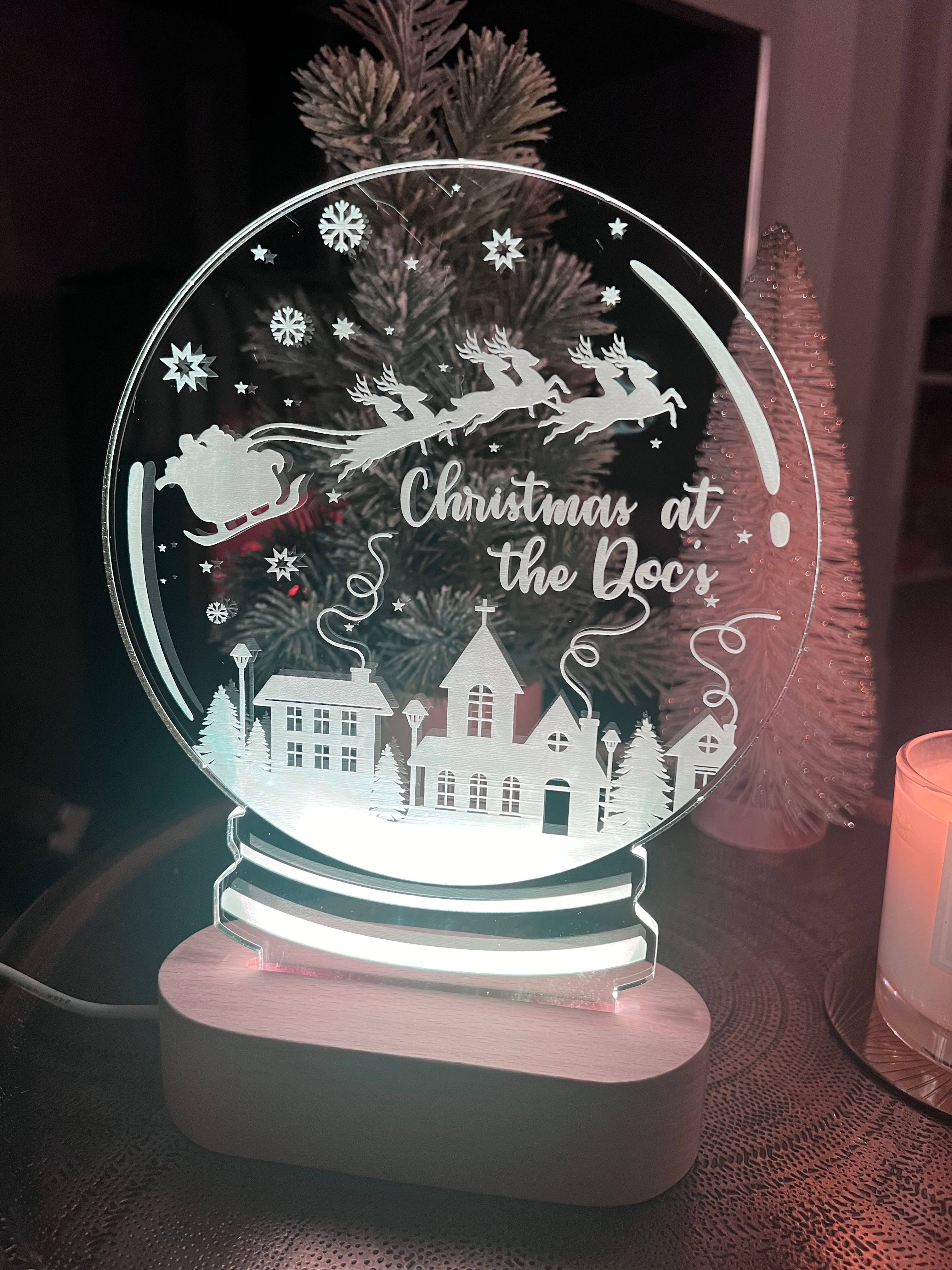 A clear acrylic Christmas snowglobe with a laser cut and engraved design featuring a reindeer, house, and Christmas tree, with a personalized text 'Christmas at the Door'.